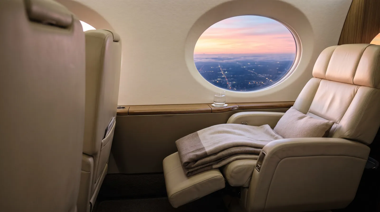 Zurückgelehnter Sitz und ein Glas Wasser am Fenster eines Business Jets bei Sonnenaufgang